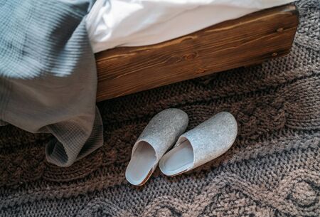 The pair of gray home slippers near the wooden bed on the knitted floor in the cozy bedroom. Home...