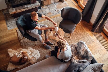 Father and son together at the home living room. Boy sitting on comfortable sofa and his daddy pouring hot tea into white cups. Beagle dog lying on sheepskin. Peaceful family moments concept image.の写真素材