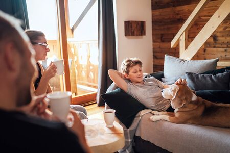 Cozy family tea time. Father, mother and son  at the home living room. Boy lying in comfortable sofa and  stroking their beagle dog and smiling. Peaceful family moments concept image.の写真素材