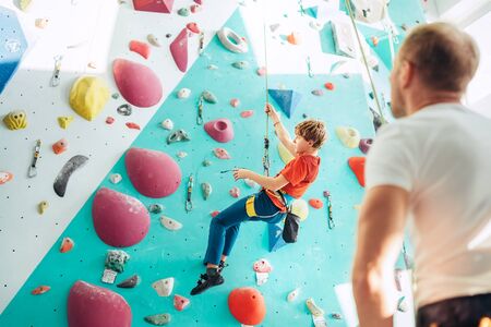 Father and teenager son at indoor climbing wall hall. Boy is hanging on the rope using a climbing harness and daddy belaying him on the floor using a belay device. Happy parenting concept image.の写真素材