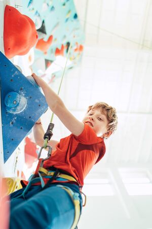 Teenager boy at indoor climbing wall hall. Boy is climbing using a top rope and climbing harness and somebody belaying him from floor. Active teenager time spending concept image.の写真素材