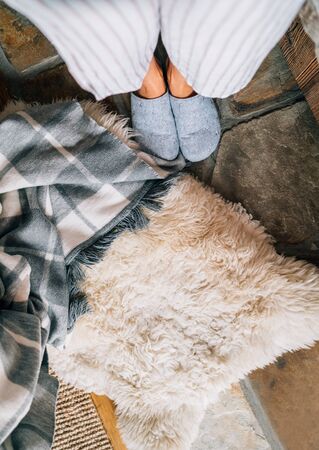 Top view of a pair of gray home slippers on female's feet near the natural white sheep sheepskin with warm plaid dropped on the stone floor in the cozy bedroom. Weekend waking up concept image.の写真素材