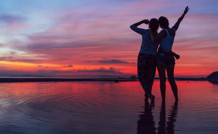 Two girlfriends hugging up and enjoying a rose/pink sunset sky on the sea beach on the Samui Island,Thailand. Calm warm countries vacation concept image.の写真素材