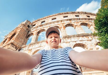 Young smiling teenager boy using two hands carrying a photo camera for taking a selfie portrait on the Pula city Amphitheatre ruins in Croatia. Famous landmarks traveling and vacation concept image.の写真素材