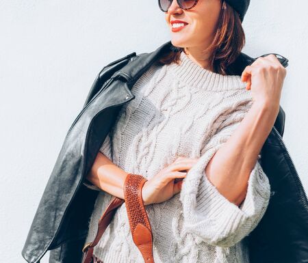 Smiling female dressed fashion style warm knitted sweater with black leather biker jacket and Beanie Hat with flap bag trendy sunglasses posing on the white wall background.の写真素材