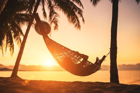 Young woman relaxing in hammock hinged between palm trees on the sand beach at orange sunrise morning timeの写真素材