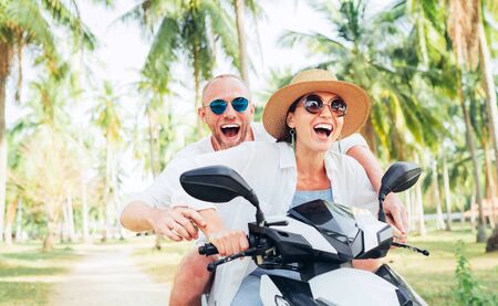 Smiling in love couple travelers riding motorbike under palm trees in their island vacationの写真素材