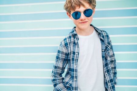 Fashion portrait of caucasian blonde hair 12 year old teenager boy dressed white t-shirt with checkered shirt in blue sunglasses with turquoise blue background wall background. Teens fashion concept.の写真素材