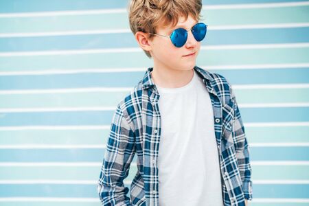 Fashion portrait of caucasian blonde hair 12 year old teenager boy dressed white t-shirt with checkered shirt in blue sunglasses with turquoise blue background wall background. Teens fashion concept.の写真素材