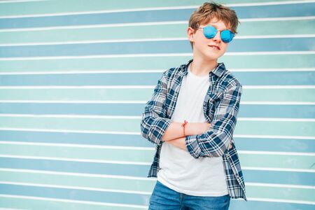 Fashion portrait of caucasian blonde hair 12 year old teenager boy dressed t-shirt and checkered shirt in blue sunglasses posing on turquoise blue background wall background. Teens fashion concept.の写真素材