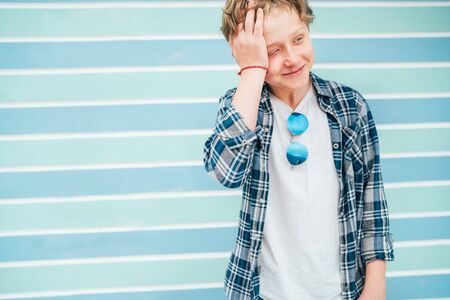 Fashion portrait of caucasian blue-eyed blonde hair 12 year old teenager boy dressed t-shirt and checkered shirt with sunglasses cramped because of shooting turquoise blue background wall backgroundの写真素材