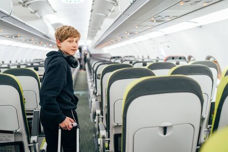 Smiling blonde hair teenager portrait staying in aircraft corridor with headphones with cabin trolley bag. Kids traveling or unaccompanied child in aircraft concept image.の写真素材