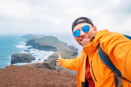Smiling brightly dressed backpacker man taking selfie portrait as he hiking and enjoying Atlantic ocean viewの写真素材