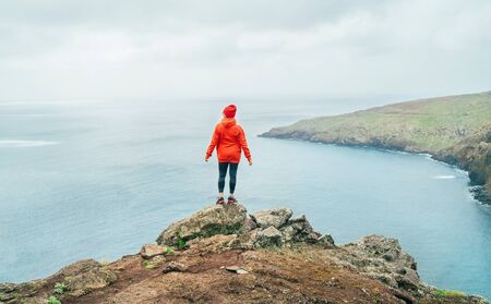 Trail runner female dressed orange sporty hoodie and red cap resting on the cliff and enjoying Atlantic ocean view on Ponta de Sao LourenÃ§o peninsula -the easternmost point of Madeira island, Portugalの写真素材