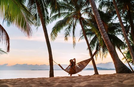 Young woman sitting in hammock swinging on the exotic island sand beach at sunrise timeの写真素材