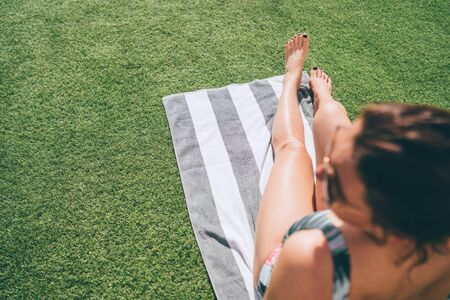 Top view of pretty female`s feet sunbathing on the stripped towel lying on green grass near swimming...