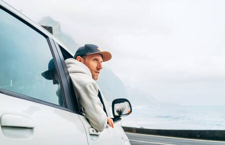 Man dressed warm hoodie and baseball cap driving his new car on the lonely road leading by the sea coastの写真素材