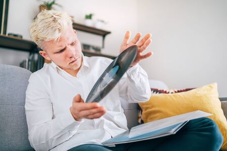 Portrait of young man sitting in comfortable sofa in living room checking out 12-inch vinyl LP recordの写真素材