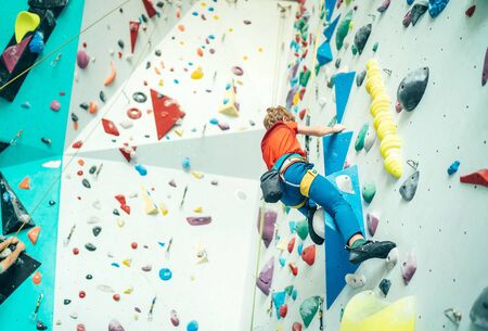 Teenager boy at indoor climbing wall hall. Boy is climbing using a top rope and climbing harness and somebody belaying him from floor. Active teenager time spending concept image.の写真素材