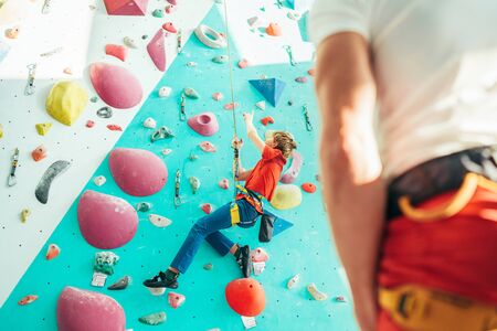 Father and teenager son at indoor climbing wall hall. Boy is hanging on the rope using a climbing harness and daddy belaying him on the floor using a belay device. Happy parenting concept image.の写真素材