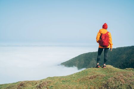 Young female backpacker dressed orange waterproof jacket hiking by the mountain above the cloud...