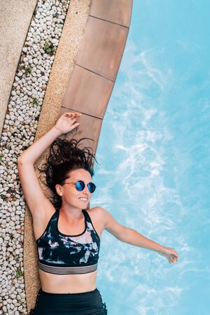 Beautiful sincerely smiling woman in blue sunglasses lying on the  swimming pool paved edgeの写真素材
