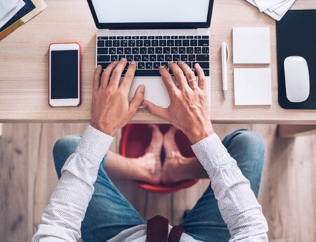  Comic modern office table top view shot. Businessman typing on laptop keyboard and soaring his feet in Foot hot Bath under table. Distance work in worldwide quarantine time concept.の写真素材