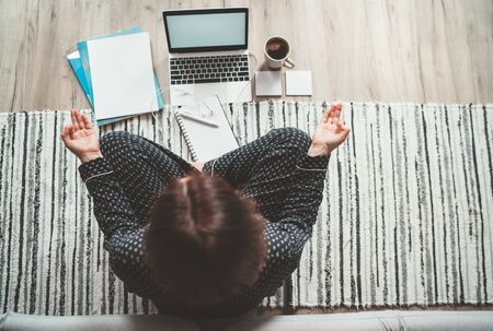  Businesswoman dressed in pajamas meditating with morning coffee with Gyan Mudra palms.On the floor office with laptop, papers top view shot. Distance work in worldwide quarantine time concept.の写真素材