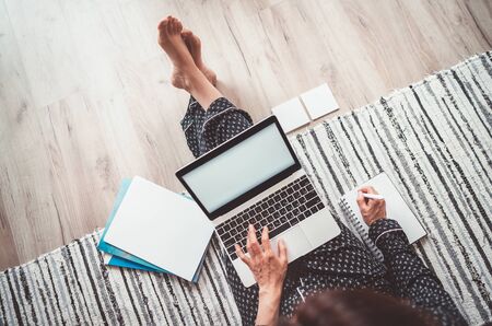 Business woman barefoot dressed pajamas writing notes sitting on living room floor office with laptopの写真素材