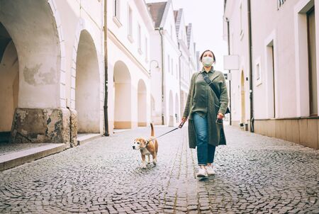 Young female fancy-dressed using a face mask as a coronavirus spreading prevention walking with her beagle dogの写真素材