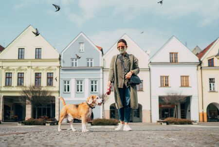 Young female fancy-dressed using a face mask as a coronavirus spreading prevention walking with her beagle dogの写真素材