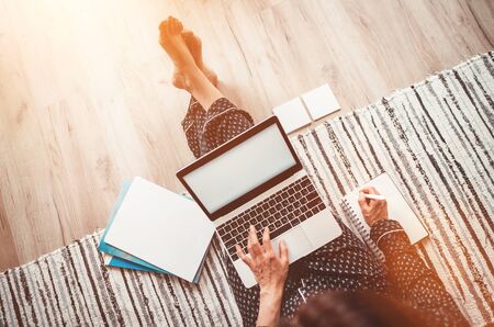  Business woman barefoot dressed pajamas writing notes sitting on living room floor office with laptop, papers and other stuff top view shot.Distance work in worldwide quarantine time conceptの写真素材