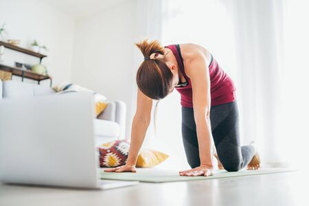 Fit sporty healthy woman on mat in Cat Majaryasana pose, doing breathing exercises, watching online yoga class on laptop computer. Healthy people and self motivation concept.の写真素材