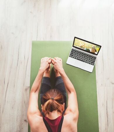 Top view at fit sporty healthy woman sitting on mat in Paschimottanasana pose, doing breathing exercises, watching online yoga class on laptop computer. Healthy people and self motivation concept.の写真素材