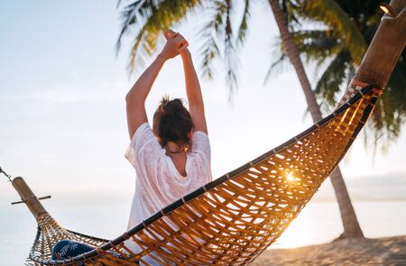 Young woman awaking and meeting morning sunrise sunlight sitting in a hammock and lazy stretching her body raising arms up on the sandy beach under the palm trees. Calm exotic places vacation conceptの写真素材