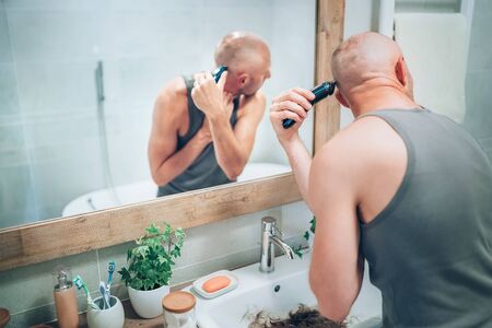 Hairless man ending shaving off all his hairs looking in bathroom mirror using an electric rechargeable Trimmer. Radical decision, image changing or body and skincare treatment concept image.の写真素材