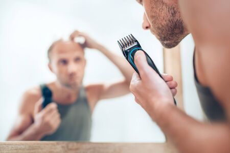Man looking at bathroom mirror and thinking about his new style haircut analyzing hairs.Electric rechargeable Trimmer close up focus image. Radical decision, life-changing or body and skincare conceptの写真素材
