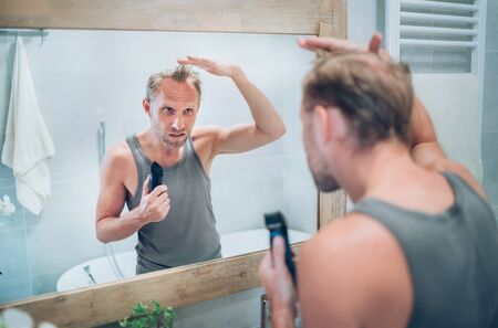 Man looking at bathroom mirror and thinking about his new style haircut analyzing hairs holding electric rechargeable Trimmer. Radical decision, life-changing or body and skincare treatment concept.の写真素材