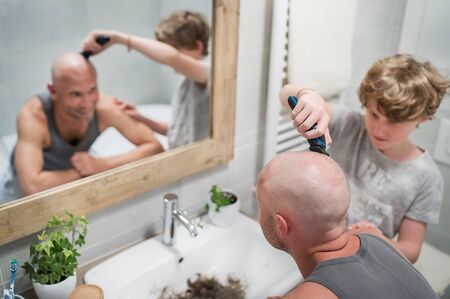 Teenager son helping his father to trim a bald head gently using electric rechargeable Trimmer in bathroom. Funny home scene and family relatives concept image. の写真素材