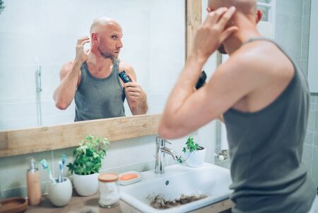 Hairless man touching his new style bald head haircut he made using an electric rechargeable Trimmer looking in bathroom mirror.の写真素材