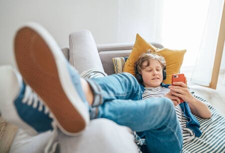 Preteen boy lying at home on cozy sofa dressed casual jeans and new sneakers listening to music and chattingの写真素材