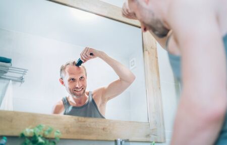 Smiling Man making new style haircut trimming a hairs using an electric rechargeable Trimmer looking...