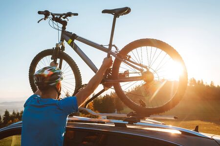 Young Man dresses modern cycling clothes and protective helmet installing his mountain bike on the car roof with sunset backlight. Active sporty people concept image.の写真素材