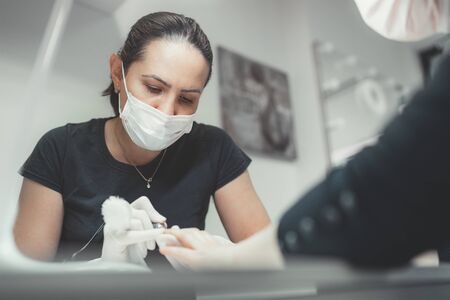 Professional manicure master dressed black in safety face mask using Electric Nail Polisher Tool for Glazing treatment manicure procedure. Small business everyday working concept.の写真素材