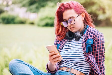 Portrait of Beautiful modern young female teenager with extraordinary hairstyle color in a checkered shirt with wireless headphones texting via smartphone. Modern teens in digital world concept imageの写真素材