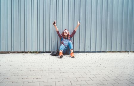 Young female teenager in a checkered shirt and jeans with headphones and smartphone sitting near a wall on the street.の写真素材