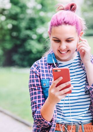 Portrait of Beautiful modern smiling young female teenager with extraordinary hairstyle color in a checkered shirt chuting via smartphone using earphones. Modern teens in digital world concept imageの写真素材