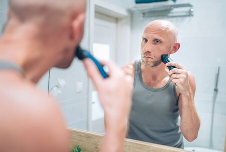 Bald man fixing his beard looking in bathroom mirror using an electric rechargeable  Trimmer.の写真素材