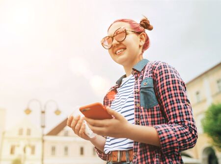 Portrait of smiling beautiful modern young female teenager with extraordinary hairstyle in checkered shirt holding a slim smartphone in hand. Modern teens or cheerful students concept image.の写真素材