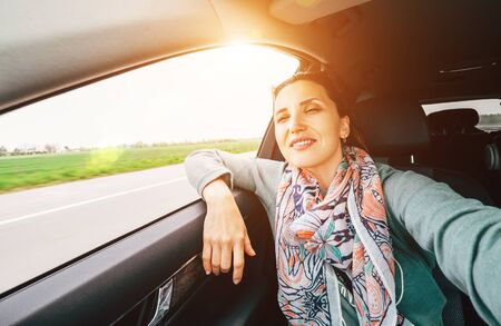 Woman making a selfie photo using smartphone and enjoying the fast car moving speed while she have a trip on the co-driver seat on the highway road. Safety car driving and people traveling concept.の写真素材